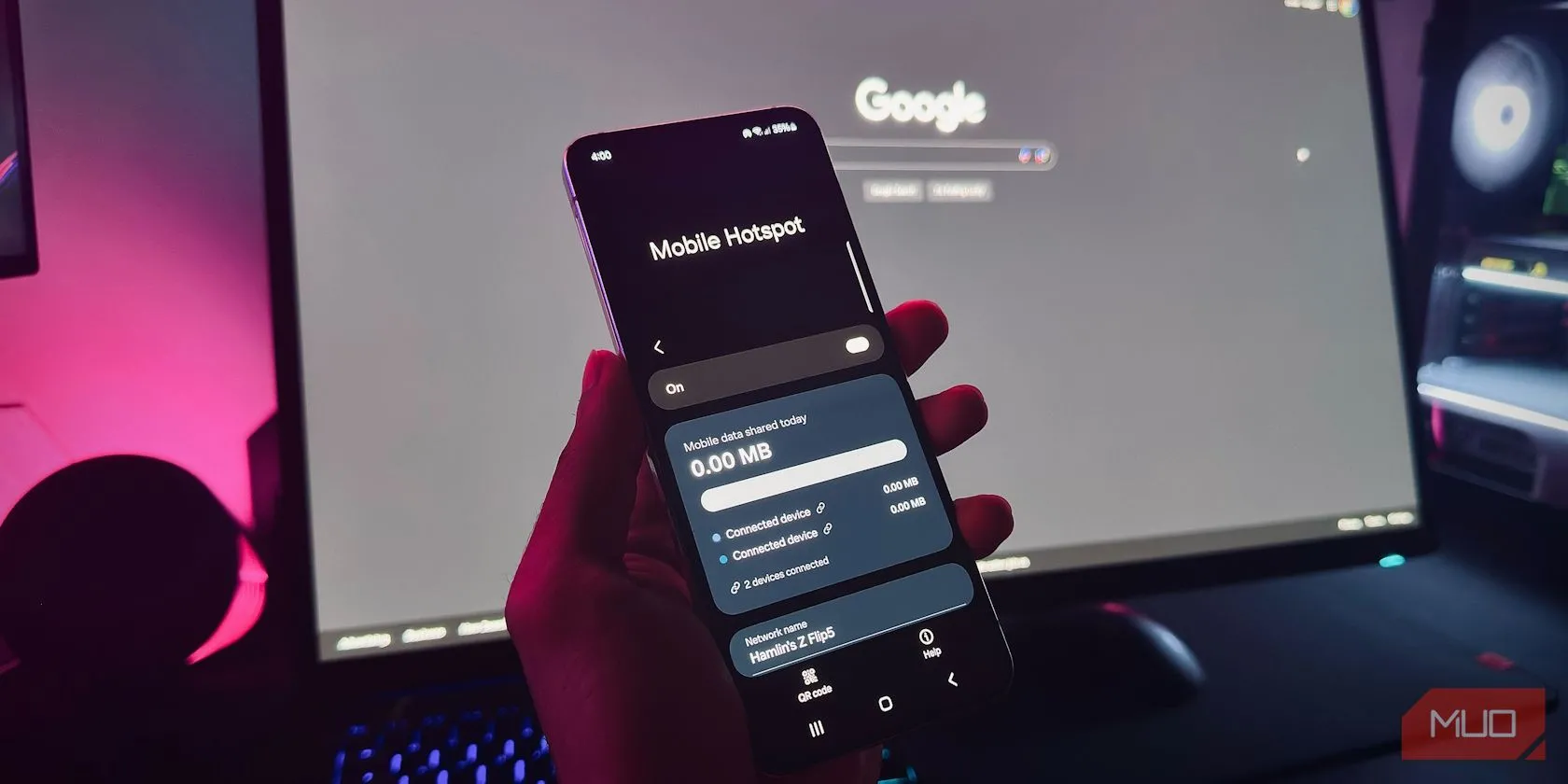 Một bàn tay đang cầm điện thoại Android hiển thị tính năng Điểm phát sóng di động trước màn hình máy tính, minh họa việc sử dụng hotspot cho công việc.